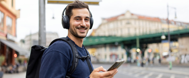 Junger Mann steht mit Kopfhörern und Handy an einer Bushaltestelle und lächelt