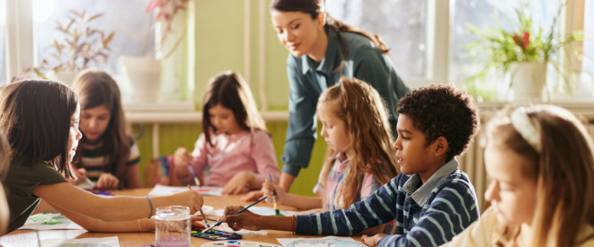 Eine Gruppe Grundschüler sitzt an einem Tisch und malt mit Wasserfarbe und die Lehrerin schaut zu