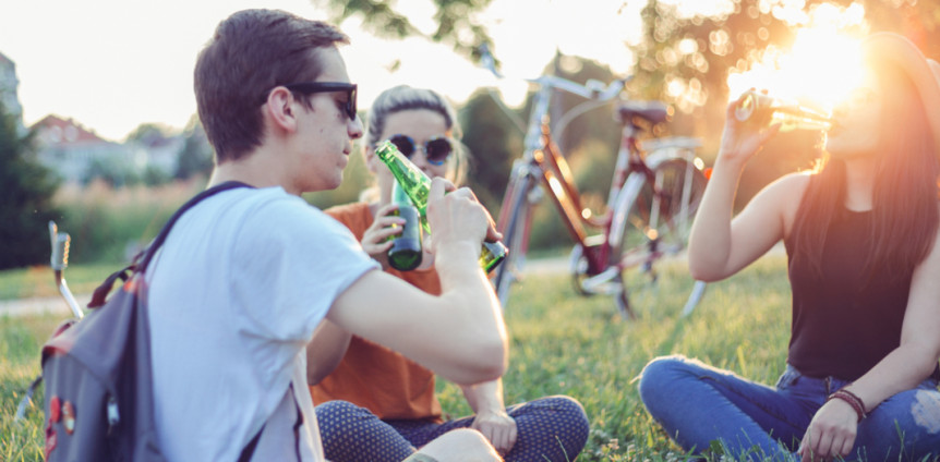Drei junge Menschen sitzen bei Sonne mit ihren Fahrrädern auf einer Wiese und trinken Bier
