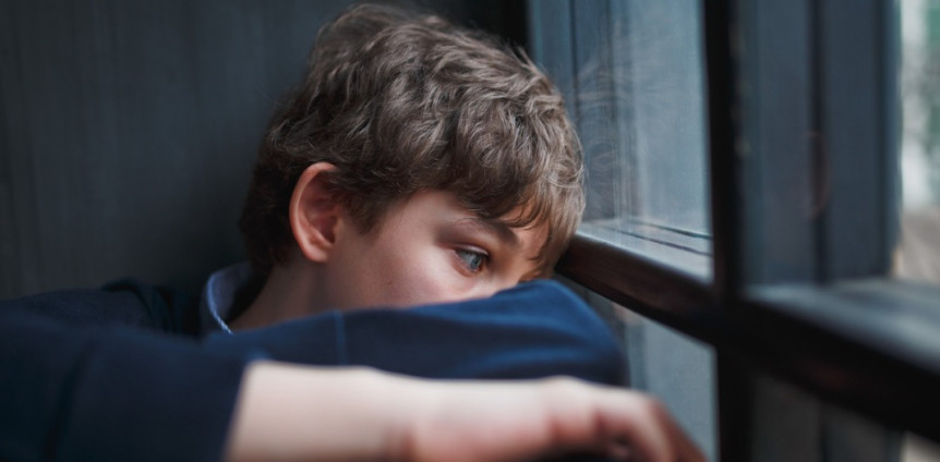 Trauriger Junge sitzt mit angezogenen Beinen am Fenster