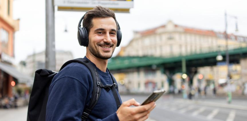 Junger Mann steht mit Kopfhörern und Handy an einer Bushaltestelle und lächelt