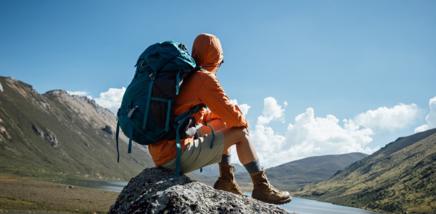 Mann in Wanderkleidung und mit Rucksack sitzt auf Stein in Gebirge und genießt Aussicht über Berge und Wasser