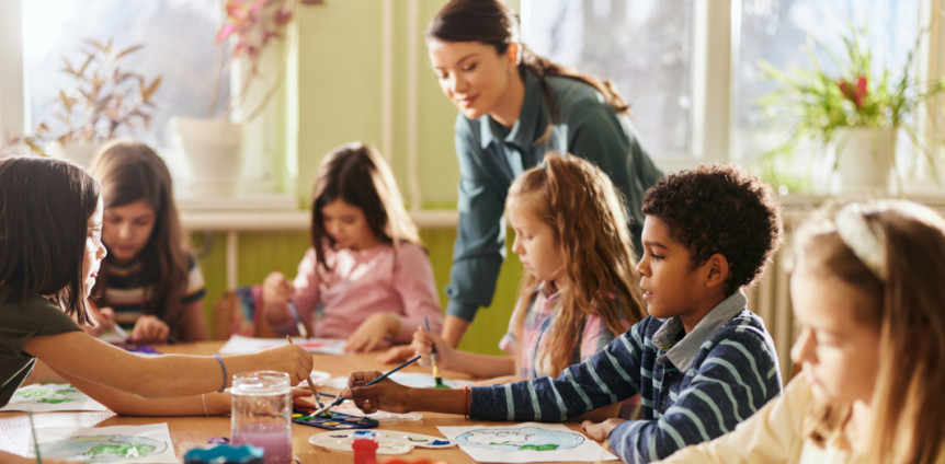 Eine Gruppe Grundschüler sitzt an einem Tisch und malt mit Wasserfarbe und die Lehrerin schaut zu