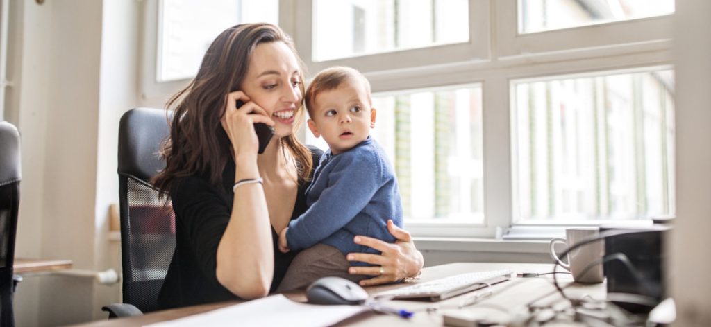 Frau sitzt mit Kleinkind im Arm am Schreibtisch im Büro und telefoniert.
