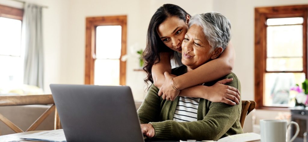 Junge Frau umarmt ältere Frau von hinten vor einem Laptop