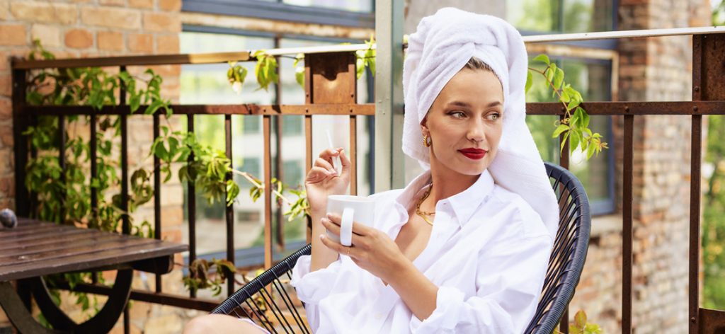 Frau mit Handtuch um den Kopf sitzt auf ihrem Balkon, trinkt Kaffee und raucht eine Zigarette.