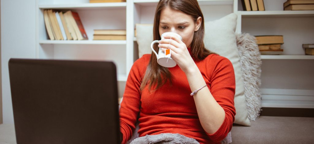 Frau sitzt vor dem Arbeitslaptop und trinkt eine Tasse Tee.