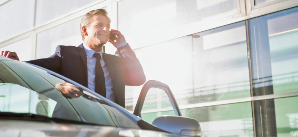 Geschäftsmann steht auf der Fahrerseite seines offenen Fahrzeugs und telefoniert am Smartphone.