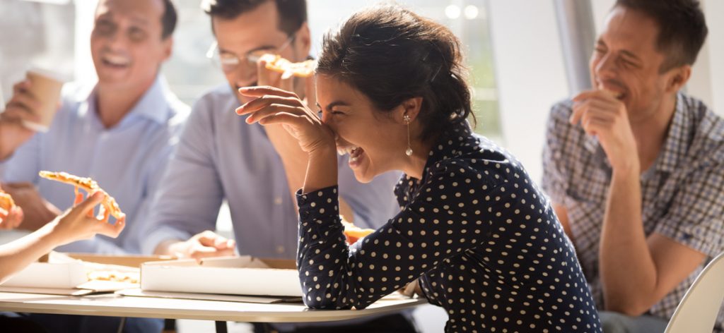 Frau lacht mit Freunden an einem Tisch. Sie essen dabei Pizza.