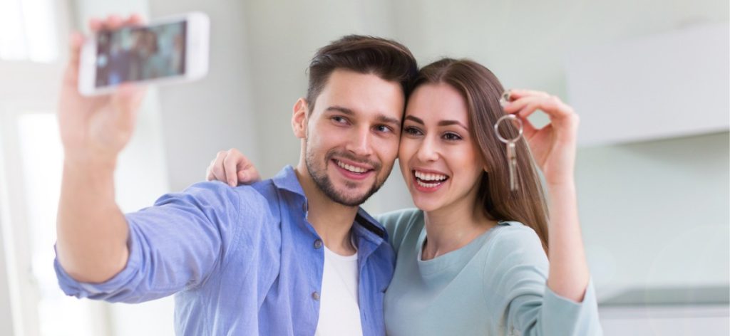 Junges Paar hält glücklich die Wohnungsschlüssel hoch und machen ein Selfie mit dem Smartphone.