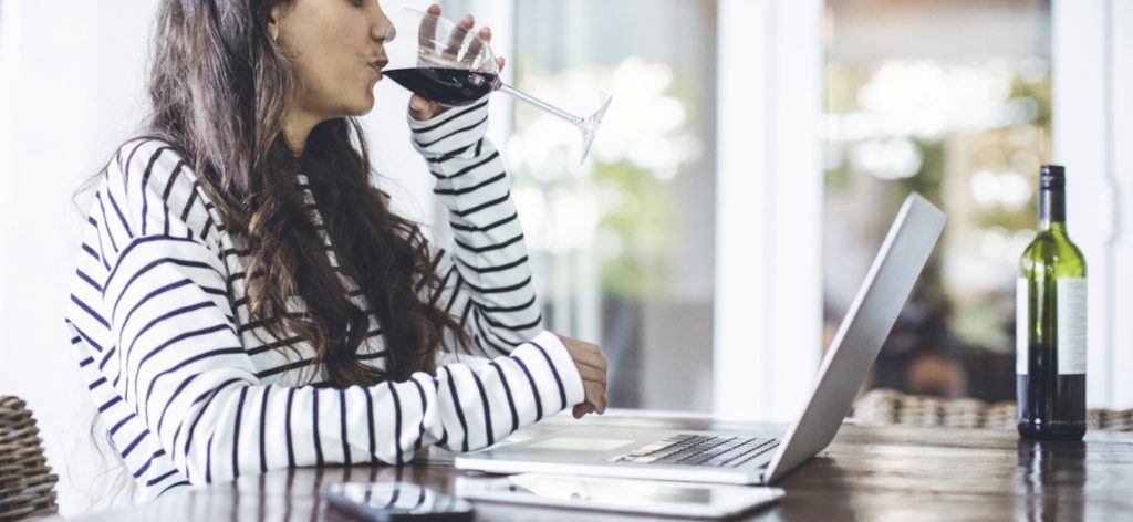 Frau tippt auf einem Laptop und trinkt gleichzeitig Wein aus einem Weinglas.