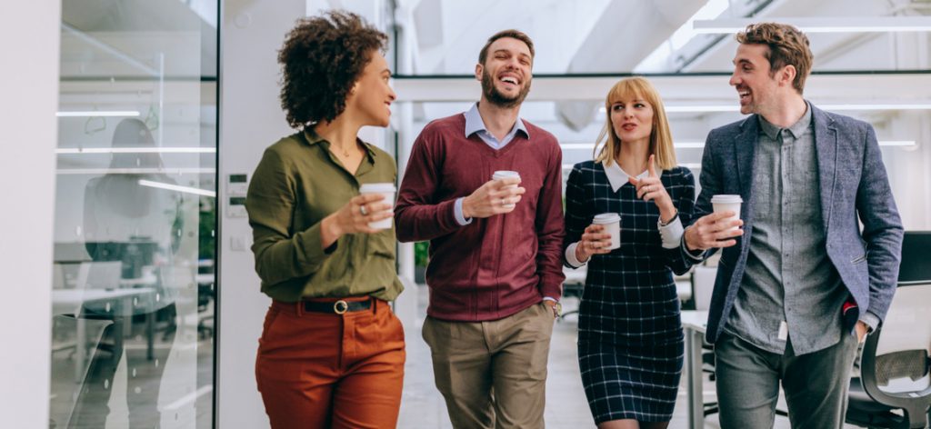 Vier Arbeitskollegen und Arbeitskolleginnen schlendern mit Kaffee in der Hand durch einen Büroflur.