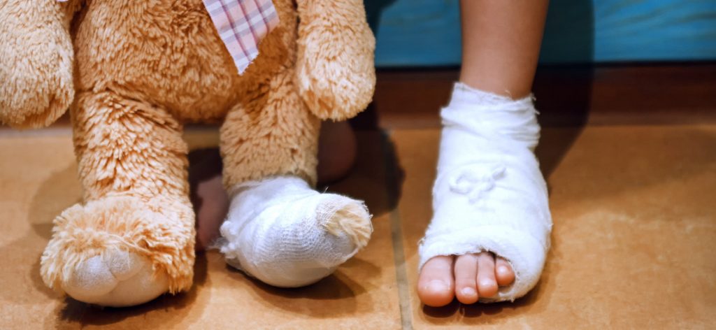 Kinderfuß mit Verband. Daneben ein Teddybein, ebenfalls mit Verband