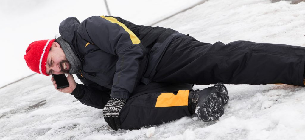 Glätteunfall auf dem Gehweg: Beweislast und Schmerzensgeld. Ein Mann liegt im Schnee, er hält sich mit deiner Hand das Knie. In der anderen Hand hält er ein Handy.