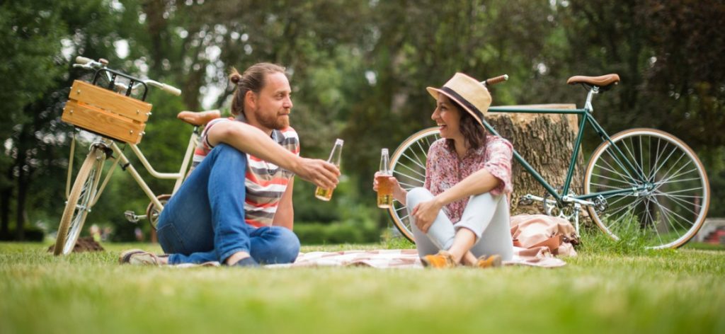 Zwei Personen sitzen auf einer Picknickdecke im Park und trinken Bier. Im Hintergrund stehen zwei Fahrräder.