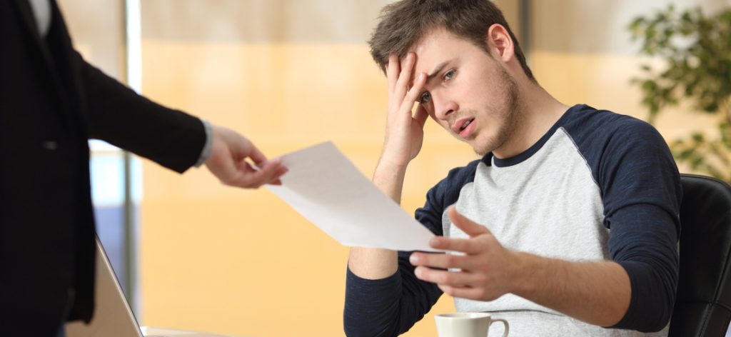 Mitarbeiter schaut entsetzt und hält eine Hand an den Kopf. Eine weitere Person gibt ihm ein Papierstück mit seiner Kündigung.