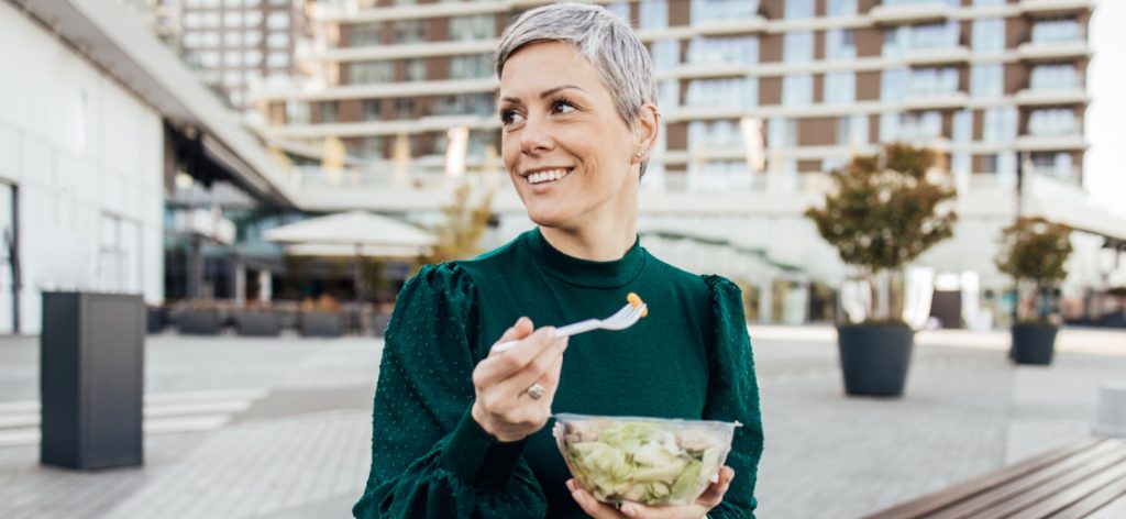 Eine junge Frau mit kurzen, grauen Haaren und grünem Oberteil hält eine Plastikschale mit Salat in der einen Hand und in der anderen Hand eine weiße Plastikgabel. Sie schaut links aus dem Bild und lächelt.