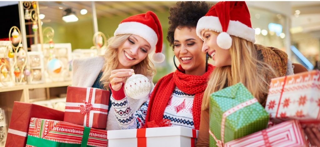 Drei Frauen mit Weihnachtsmützen und Geschenken betrachten fröhlich eine Weihnachtskugel.
