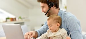 Man sitzt mit Headset vor dem Laptop und hat ein Baby auf dem Schoß