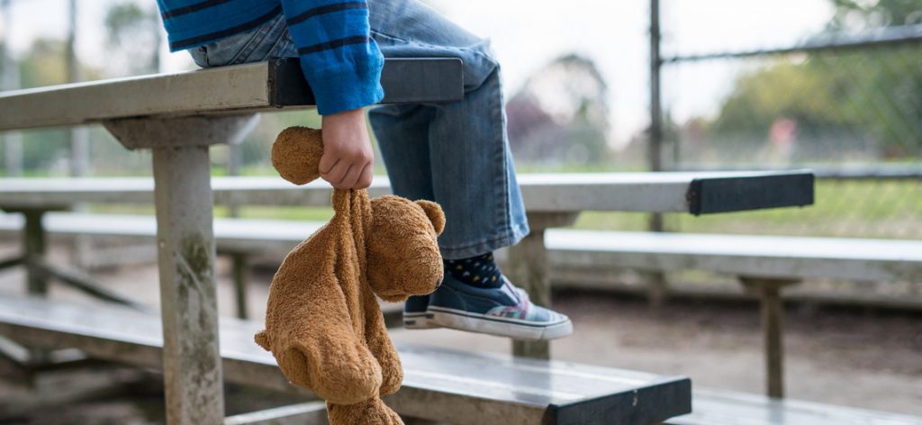 Kind sitzt mit Teddybär auf einer Sportplatzbank.