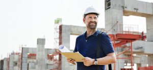 Mann mit Bauarbeiterhelm und Unterlagen steht an einer Baustelle für ein Gebäude.
