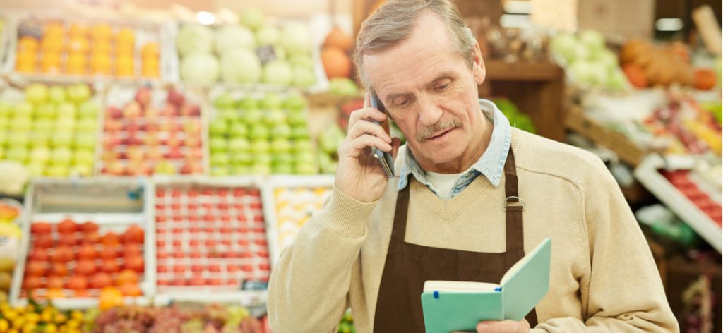 Älterer Mann telefoniert im Supermarkt und schaut auf ein Notizbuch.