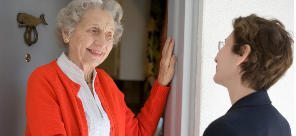 Ältere Frau mit roter Jacke steht in der Tür und unterhält sich mit einer jüngeren Frau.