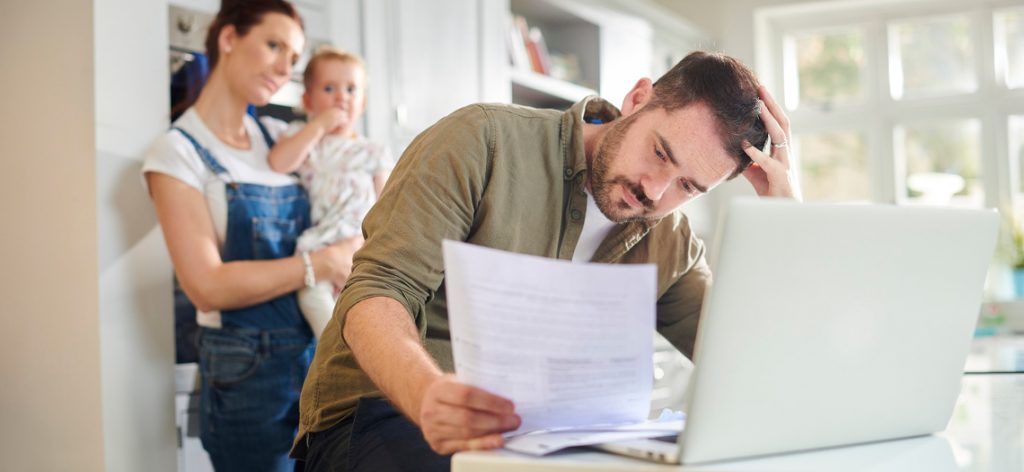 Pflicht zu Elternunterhalt: Wer muss wann zahlen? Vater sitzt verzweifelt vor Laptop und schaut auf Unterlagen. Im Hintergrund sind Frau und Kind zu sehen.