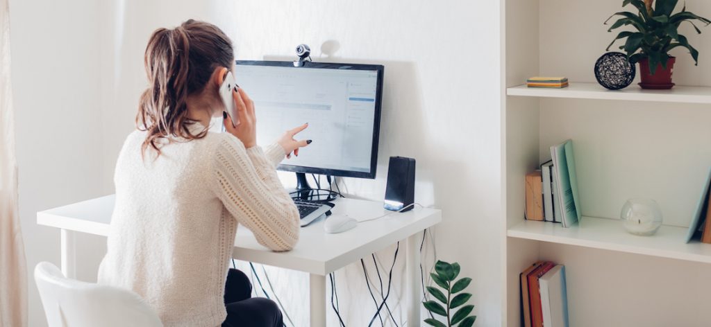 Mitarbeiterin arbeitet an ihrem PC von zu Hause und telefoniert dabei am Handy.