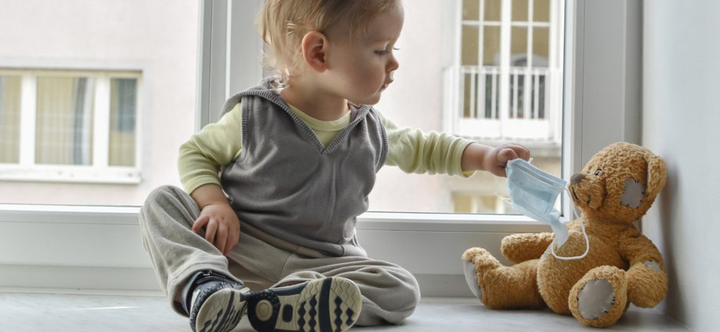 Kind in Quarantäne zieht dem Teddybär eine medizinische Maske an.