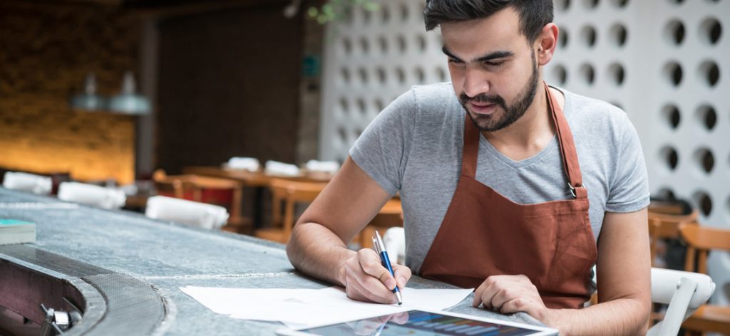 Mann mit Schürze sitzt an Bar und notiert etwas auf Papier.