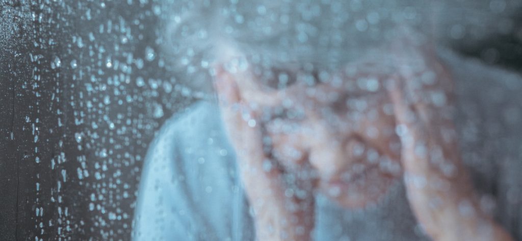 Verzweifelte alte Frau hinter einer Fensterscheibe im Regen