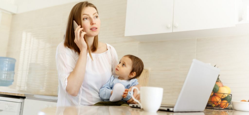 Junge Arbeitnehmerin mit Baby auf dem Arm telefoniert vor ihrem Laptop.