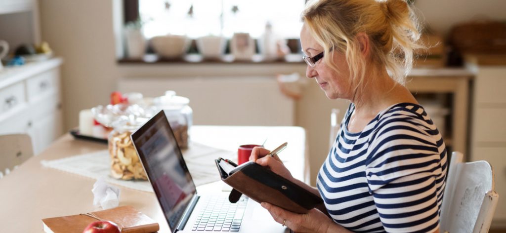 Arbeitnehmerin sitzt mit ihrem Laptop in ihrer Küche am Esstisch und schreibt in ihren Taschenkalender.