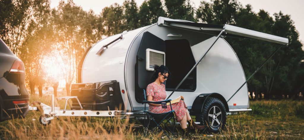 Junge Frau im Sommerkleid sitzt bei untergehender Sonne in einem Campingstuhl vor einem Wohnwagen
