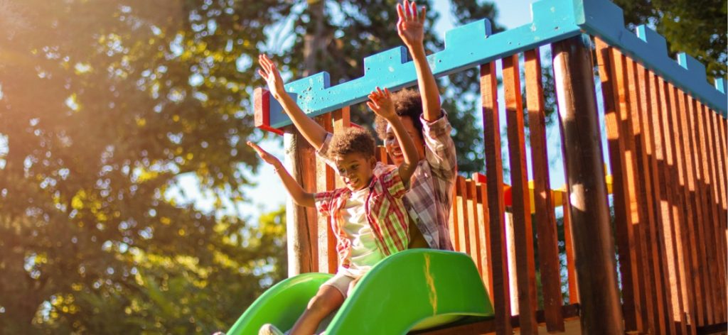 zwei Kinder sitzen oben auf einer Rutsche und halten die Hände in die Luft