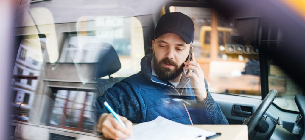 Kurierfahrer mit Cap sitzt im Auto und telefoniert und schreibt etwas auf