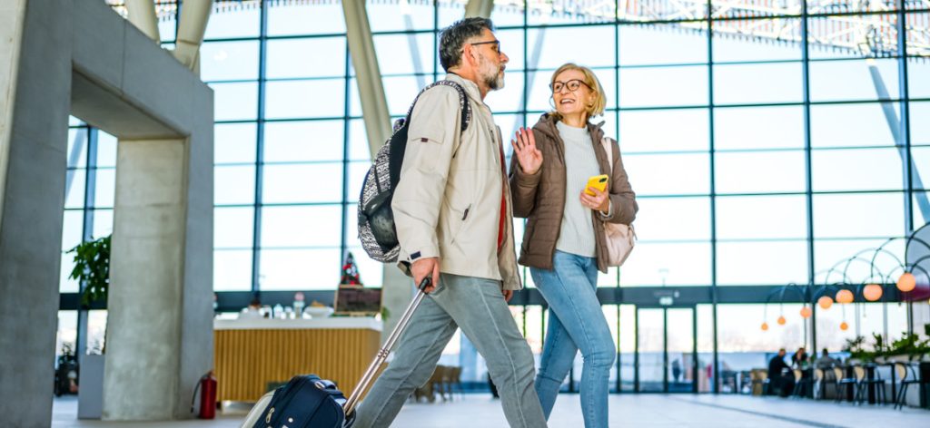 ein mittelaltes Paar läuft mit Handgepäck lächelnd durch einen Flughafen