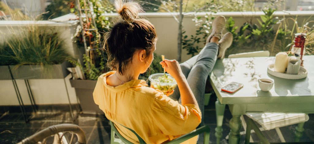 Frau sitzt in der Sonne am Tisch auf ihrem Balkon und isst einen Salat