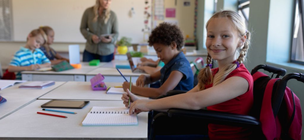 Grinsendes Mädchen im Rollstuhl mit weiteren Schülern und Schülerinnen uns Lehrerin in einem Klassenzimmer
