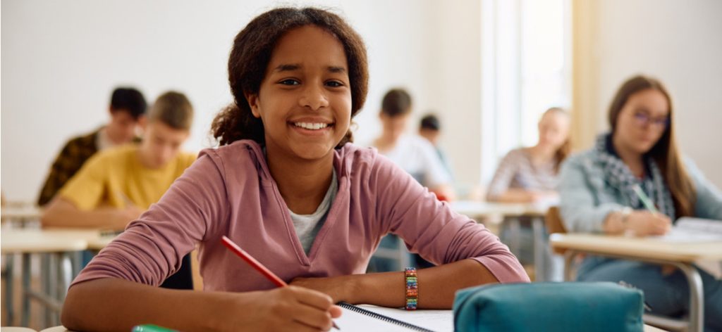 Mädchen sitzt lächelnd im Klassenraum in der Schule und schreibt in ein Heft