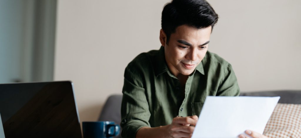 Junger Mann sitzt mit Bewerbungsunterlagen und Laptop auf dem Sofa