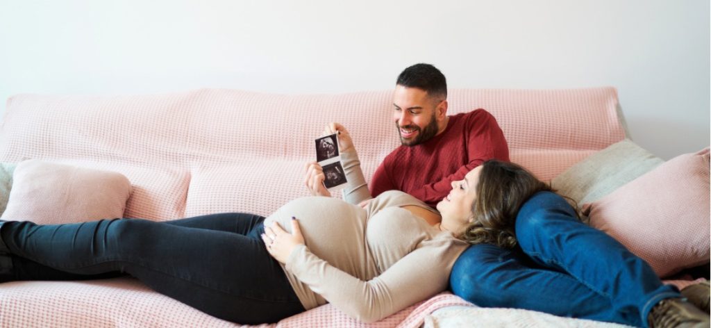 Schwangere Frau liegt auf Sofa auf Schoß des Partners und sie schauen Ultraschallbilder von Baby an