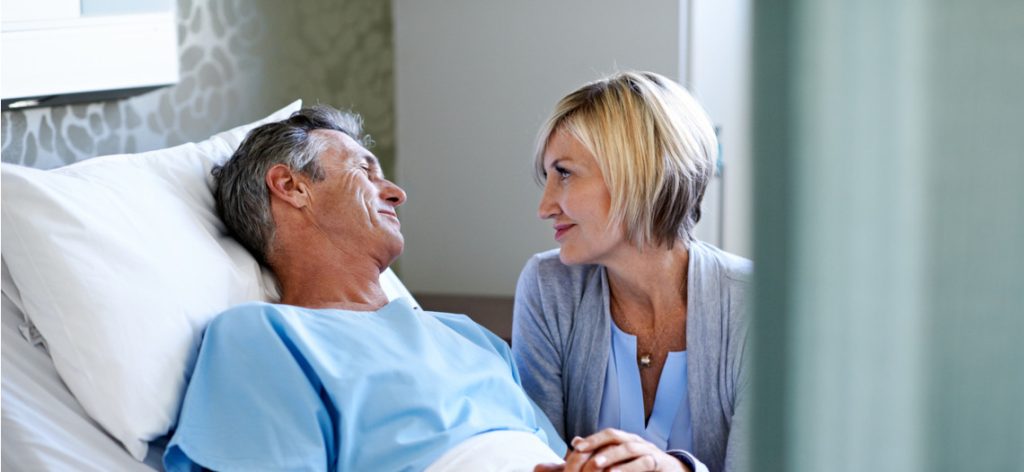 Mann in Krankenhausbett und Frau sitzt daneben und hält seine Hand