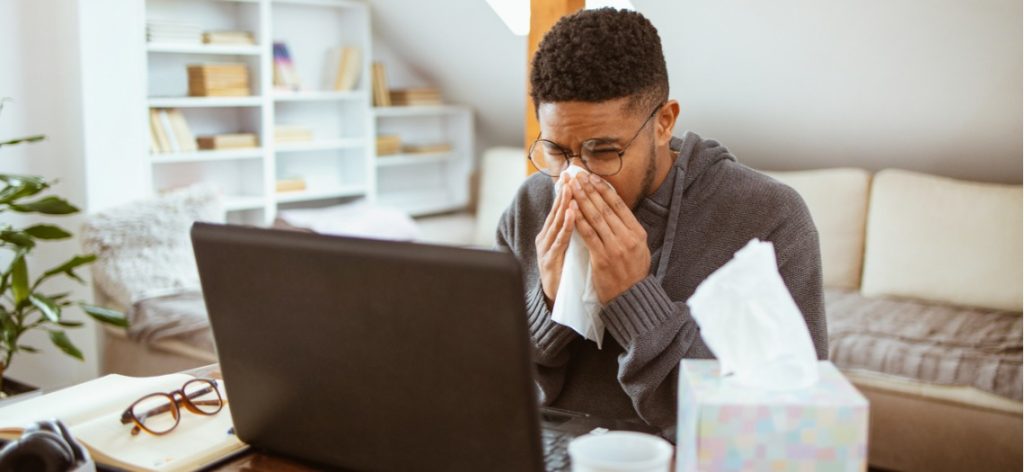 Junger Mann sitzt krank vorm Laptop am Schreibtisch und putzt sich die Nase