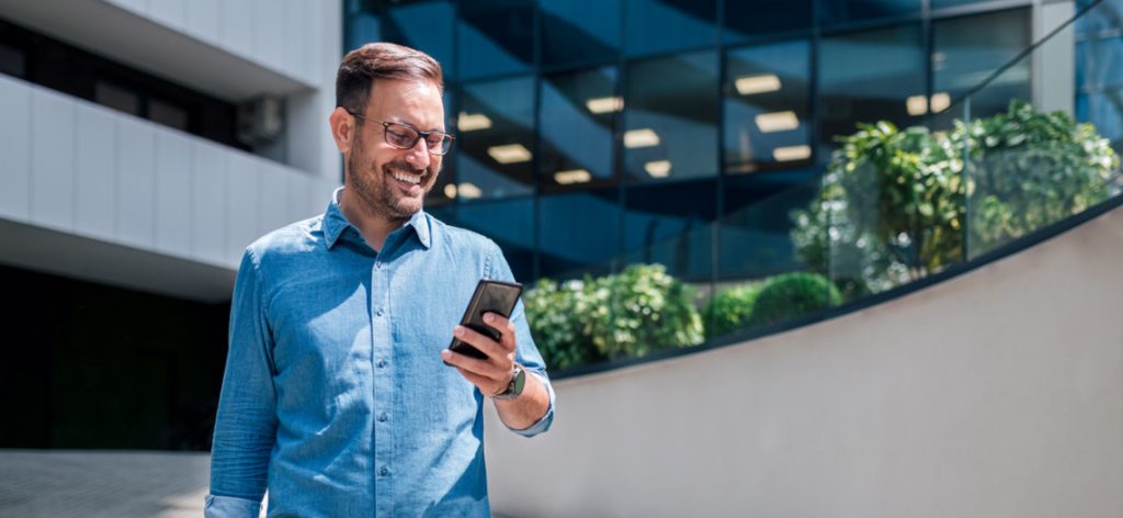 Mann vor Bürogebäude in der Sonne schaut lachend auf sein Handy