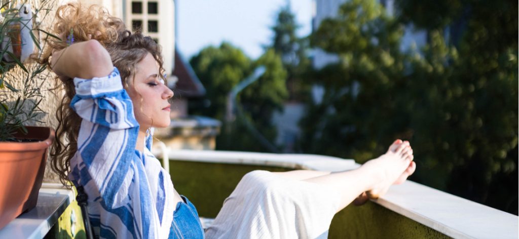 Junge Frau sitzt auf dem Balkon in der Sonne und legt die Beine hoch