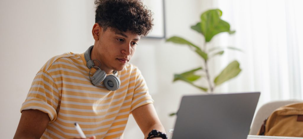 Junger Mann mit gestreiftem T-Shirt und Kopfhörern sitzt vor einem Laptop und macht Notizen