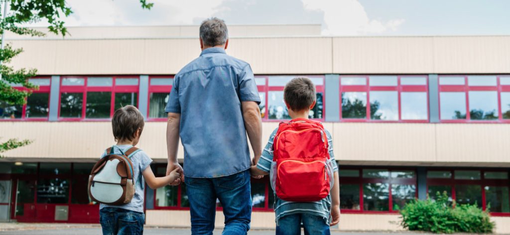Vater mit zwei Söhnen mit Rucksack stehen vor Schulgebäude