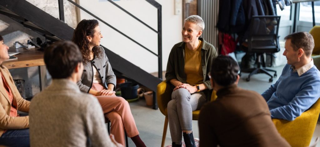 Grupp Frauen und Männer sitzt im Stuhlkreis in einer Wohnung zusammen und lacht